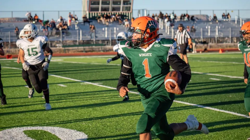 A football player in a green and orange uniform runs down the field holding a football.