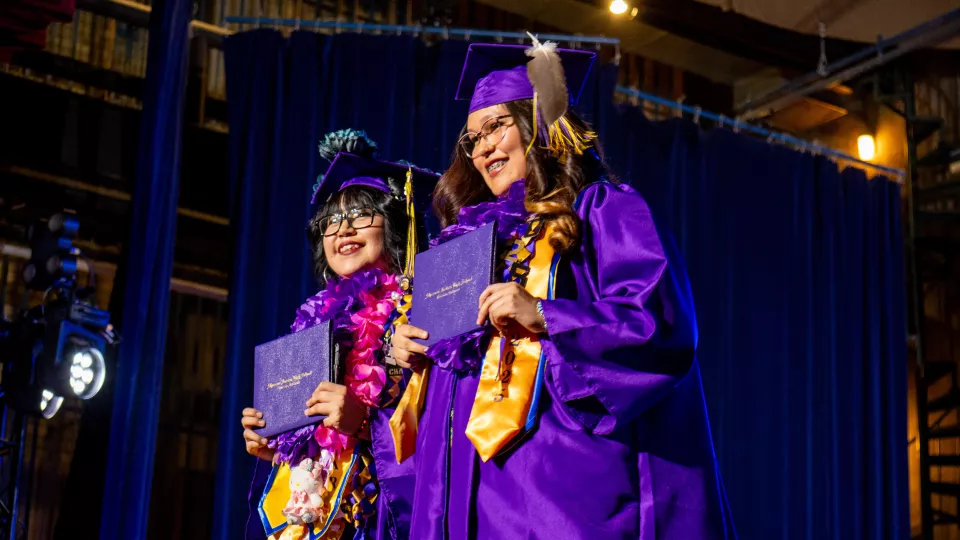 Two students in purple graduation robes accept their diplomas on stage.
