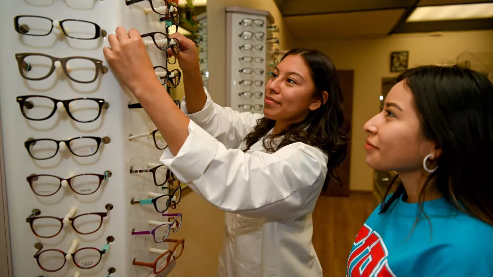 Two women stand in front of a wall display full of eyeglasses as one reaches up to pull a pair of glasses off the rack.