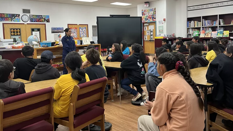 A teachers stands next to at TV at the front of a class of students all turned toward her..