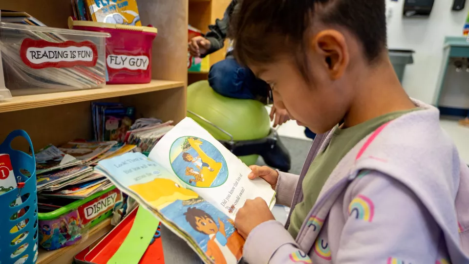 A student sits in front of a bookcase and reads a picture book.