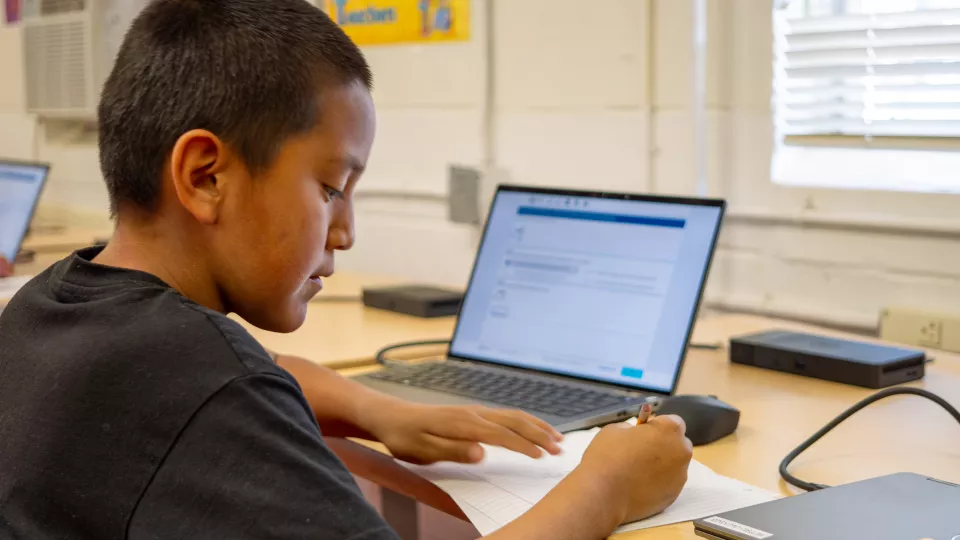 A student completes school work using both pencil and paper and a laptop.