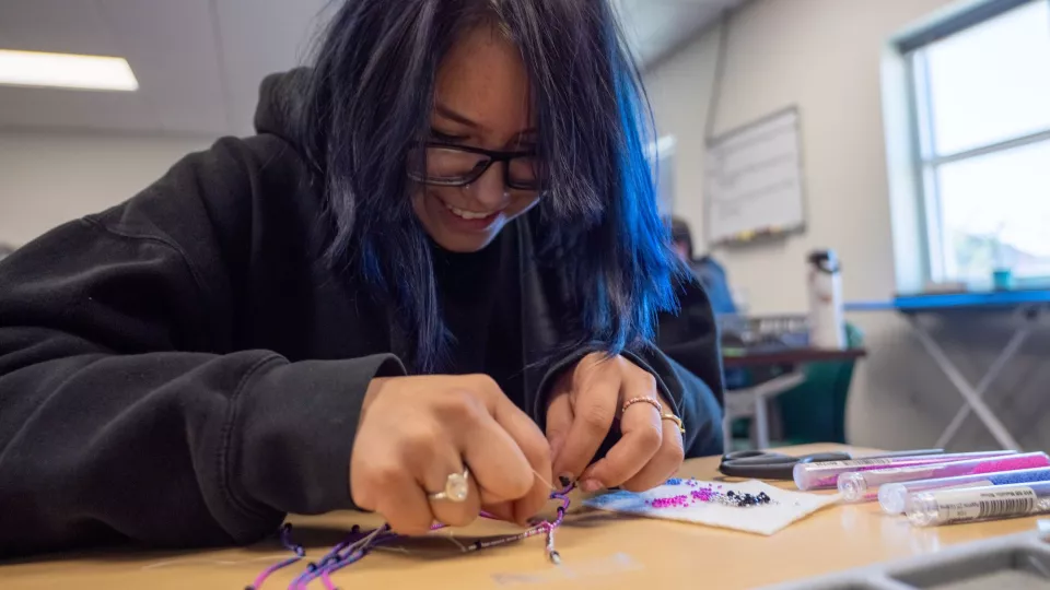 A student sits at a desk with tubes of colorful beads and works on a beading project.