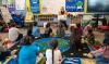 A group of students listen to their teacher read them a book while sitting on the floor.